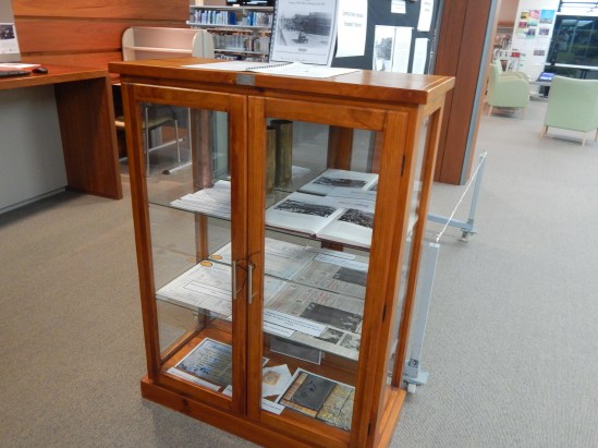 Display case containing items on loan courtesy of Parkes Historical Society. Inside are two artillery shells (one engraved in France during First World War), a copy of Ian Chambers' book Parkes: A Photographic History (belongs to Parkes Shire Library), newspaper clippings, War Service records and an Anzac biscuit and recipes.