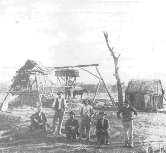 Ian Chambers noted that this photograph is of Dayspring Mine at Currajong (now called Parkes) in 1890. Owner Owen J Howard is sitting down at left with some of the miners (names unknown). This photograph shows in detail the above-ground methods employed in operating a substantial mine at that time, with good examples of the bush carpenter's trade. In the background the whim, or vertical capstan-like drum, is turned by the horse harnessed to the whim-pole. Wound around the drum is a cable which extends across to a pulley positioned above the mine and then down the shaft. By reversing the direction of the horses's circular path, the whim is thus used to raise or lower workmen, equipment and wash-dirt or ore. Photo by Harry McDade of Parkes. Source: Ian Chambers (1988) p.36